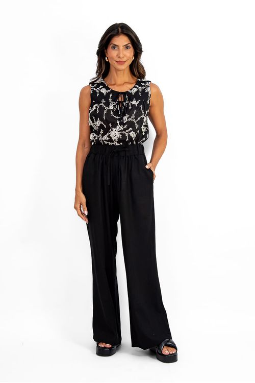 Imagem de uma mulher vestindo uma calça preta reta com cós elástico e amarração, combinada com uma blusa estampada sem mangas.