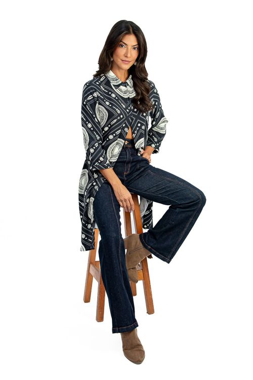 Mulher sentada em banco usando chemise longuete em viscose com estampa geométrica de esferas preto e branco, abotoada parcialmente sobre calça jeans flare escura e botinha marrom — look casual-elegante.