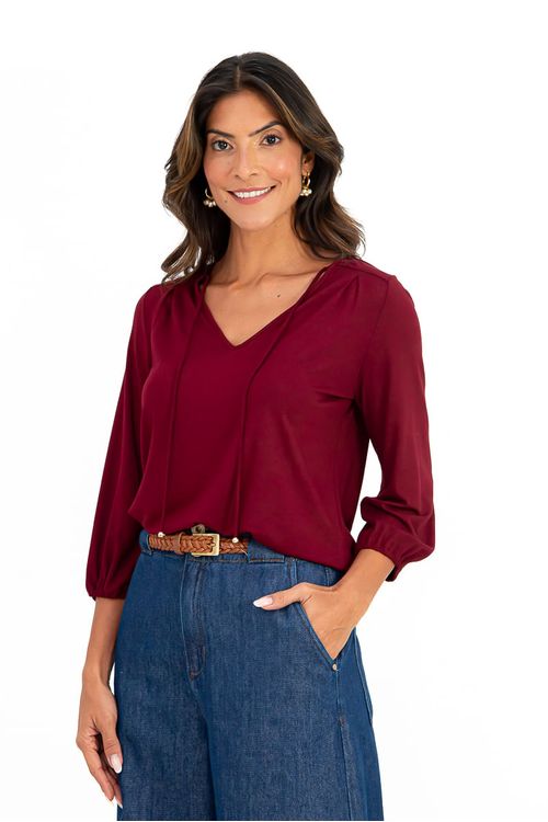 Close meio-corpo da blusa Ipanema bordô em malha viscose, destaque para decote em V com rolotê, pregas no ombro e manga 3/4 bufante com punho em elástico, estúdio fundo branco