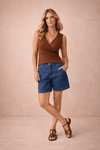 Vista frontal de uma blusa feminina transpassada chocolate, sem mangas e com franzido nos ombros, combinada com shorts jeans azuis.