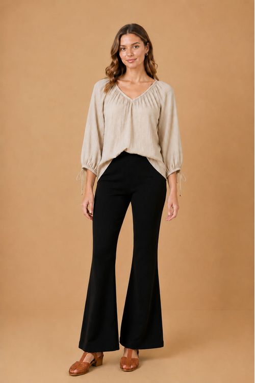 Modelo vestindo calça flare canelada preta em malha Microbaby, cós elástico, ajuste até o joelho e abertura flare na barra; look completo com blusa bege de manga 3/4 e sandália marrom de salto bloco - calça flare feminina canelada preta